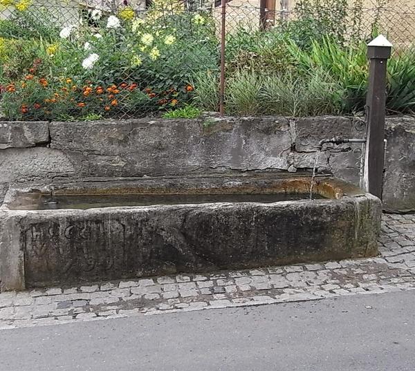 Dorfbrunnen mit Brunnenstock aus Holz
Auch hier steht ein Brunnen der schon Jahrhunderte überdauert hat. Der Brunnenkasten, so nennt man hier das Brunnen-Becken, ist noch aus Naturstein gefertigt.